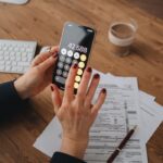 Hands using smartphone calculator over documents on a wooden table, indicating financial calculation or budgeting.